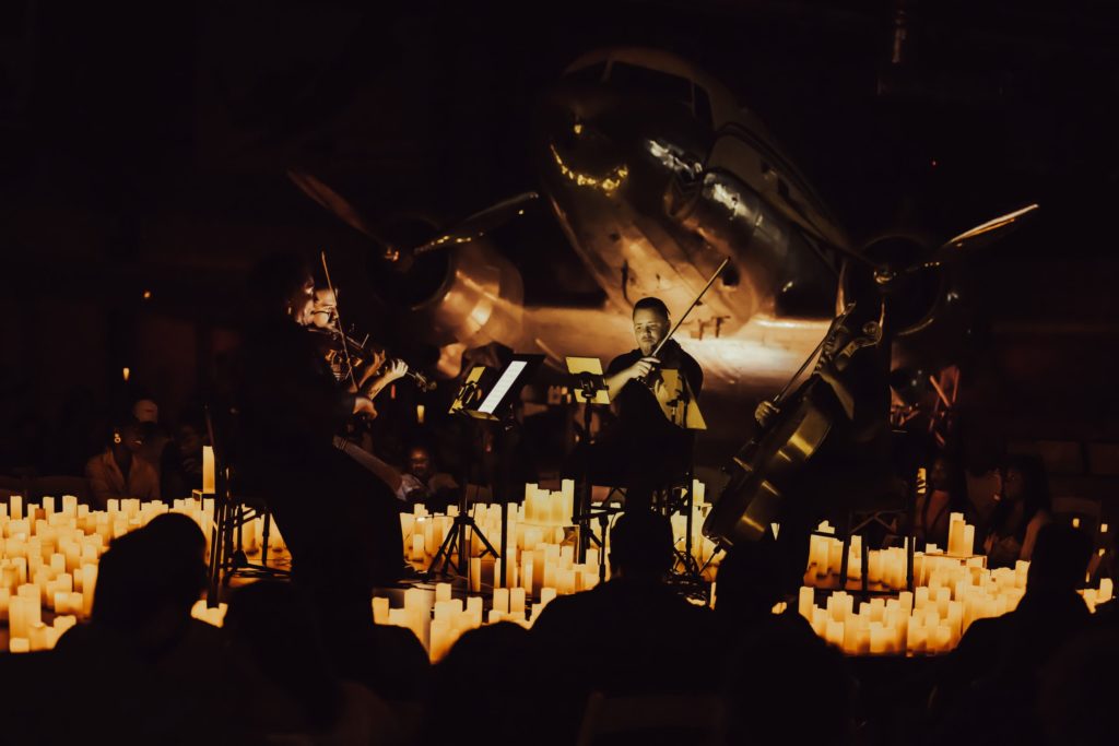The silhouette of an audience is facing a string quartet performing on a stage surrounded by candles and an aircraft imposing on the image.